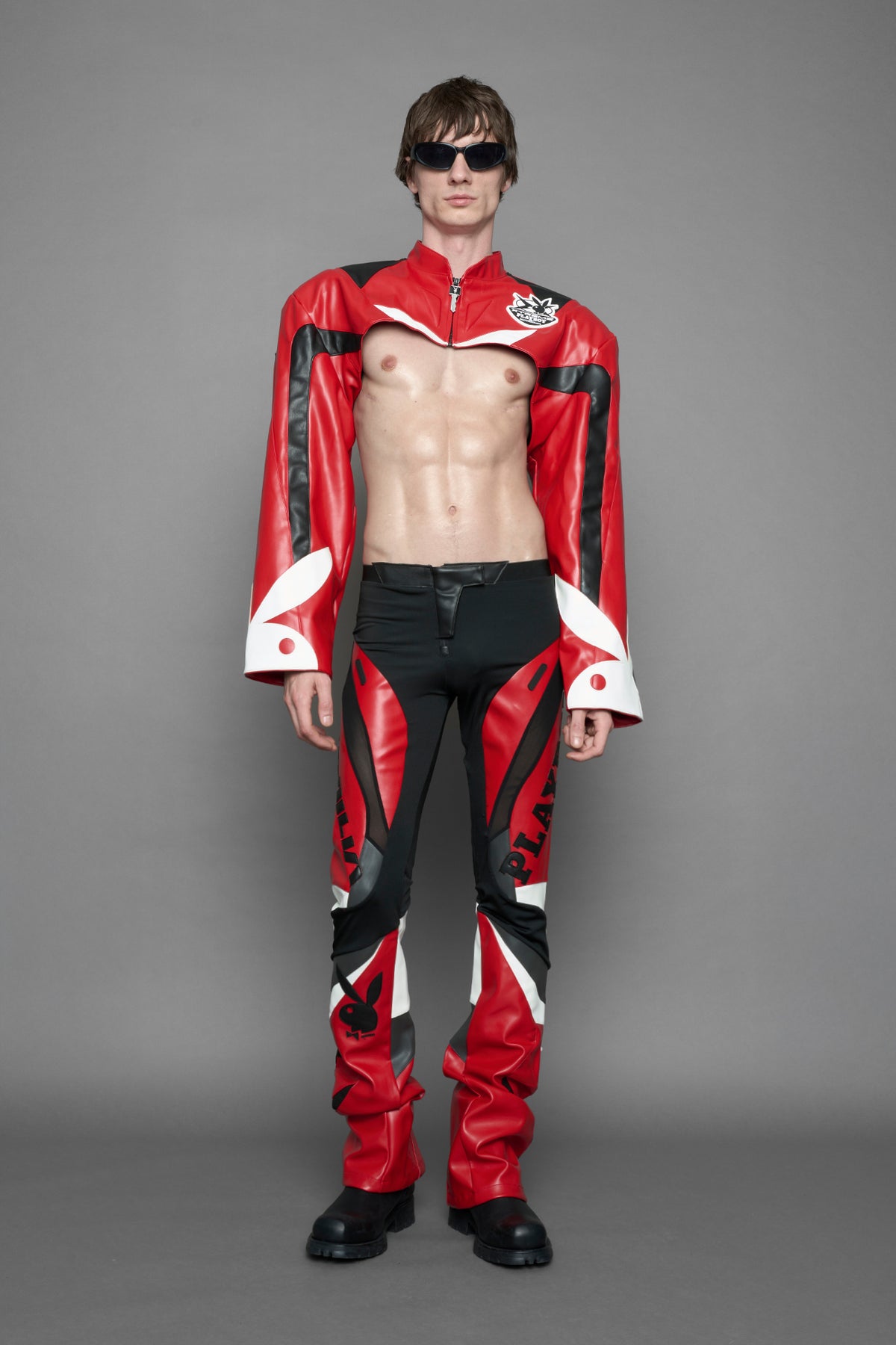 model wearing a vegan leather cropped jacket with broad shoulders in red with black and white paneling. The jacket has PU leather embroidery of 'Namilia' and 'Playboy' with a key formed zipper pull and a rubbertag on the left breast.