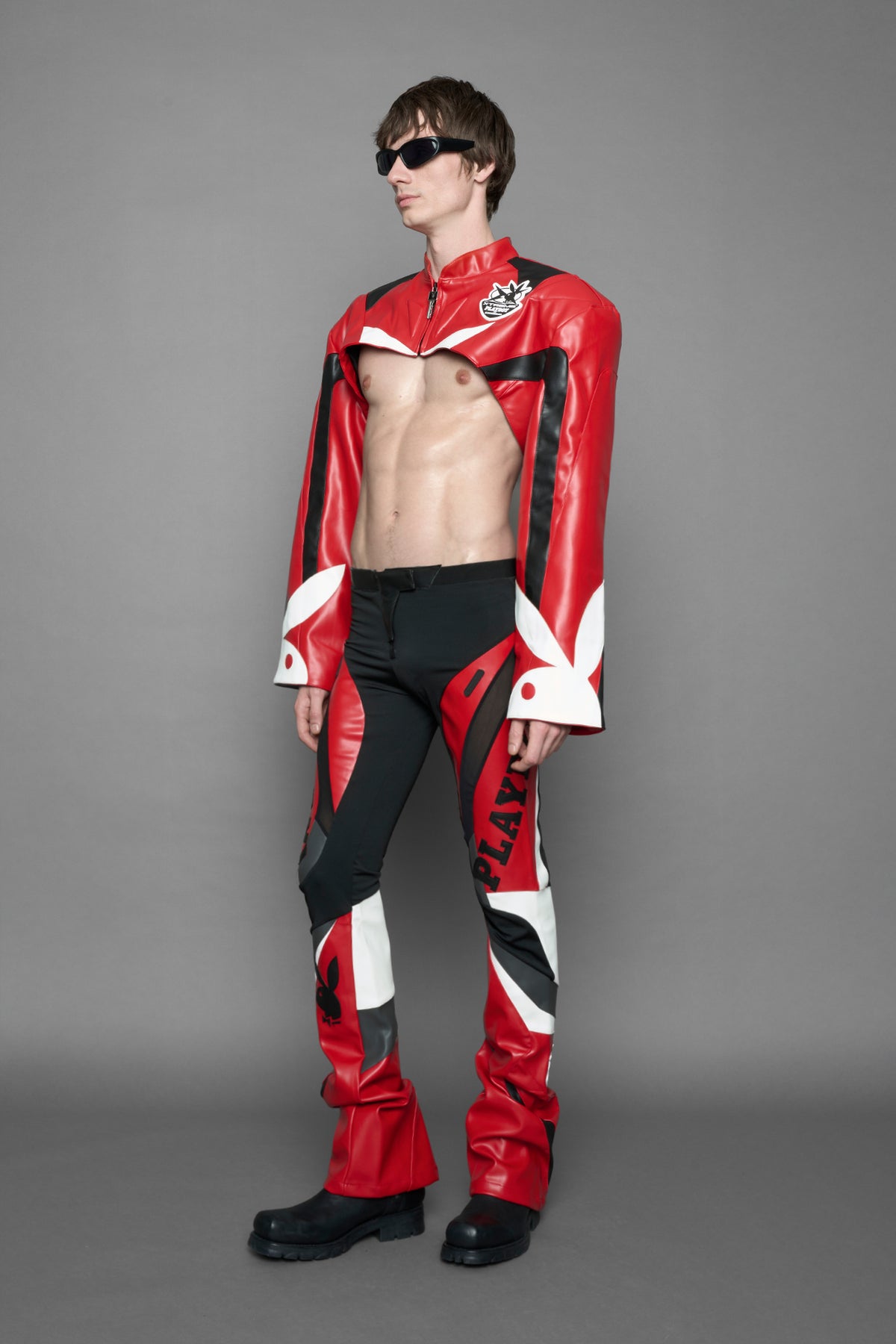 model wearing a vegan leather cropped jacket with broad shoulders in red with black and white paneling. The jacket has PU leather embroidery of 'Namilia' and 'Playboy' with a key formed zipper pull and a rubbertag on the left breast.