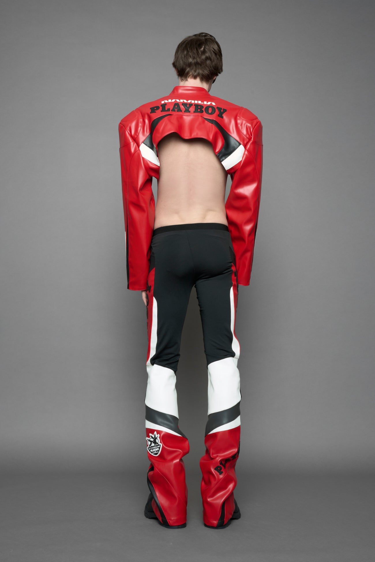 model wearing a vegan leather cropped jacket with broad shoulders in red with black and white paneling. The jacket has PU leather embroidery of 'Namilia' and 'Playboy' with a key formed zipper pull and a rubbertag on the left breast.