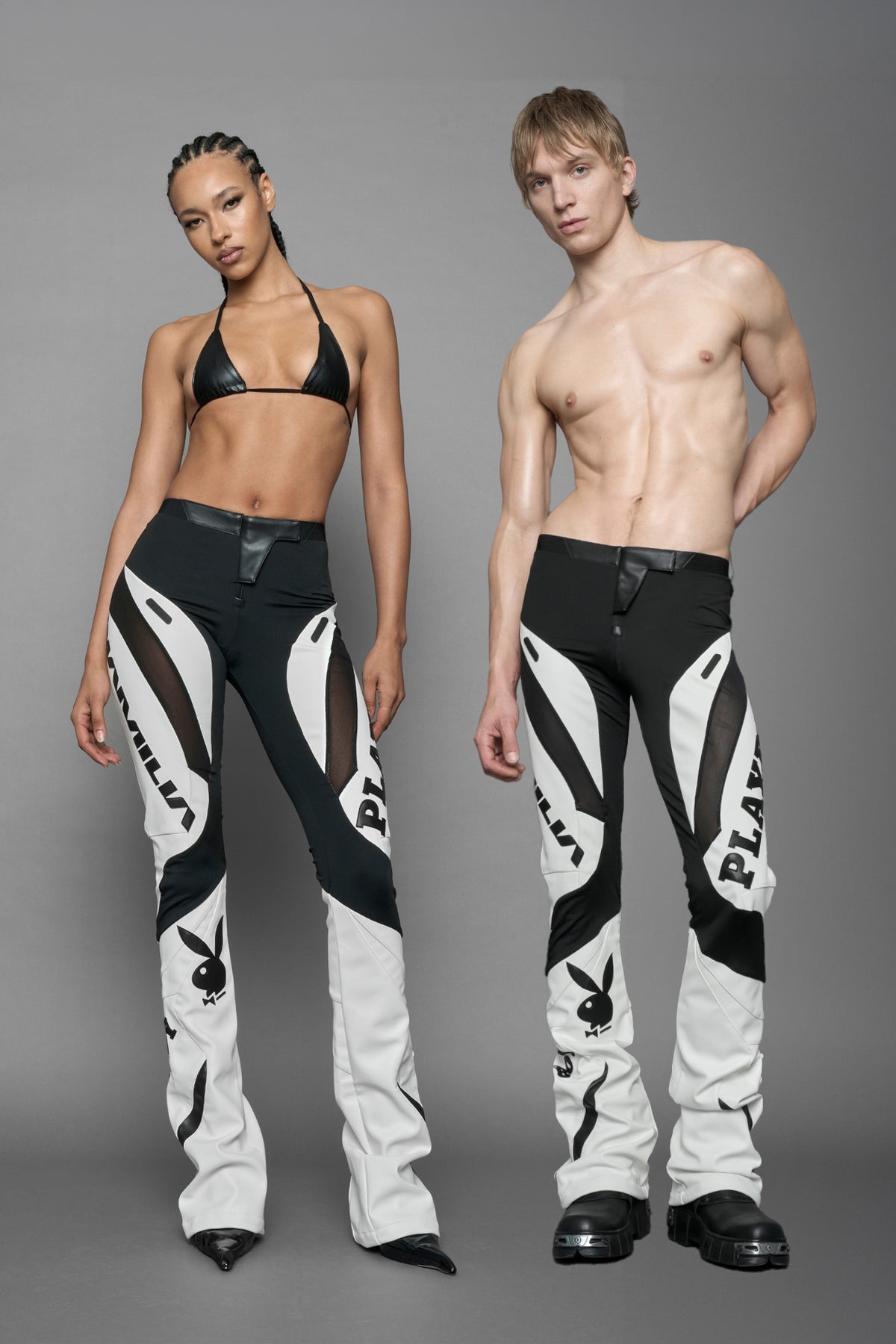 model wearing a white and black form fitted Pants with PU Leather Embroidery of 'Namilia', 'Playboy'. The pants have black Mesh fabric panels and are slightly flared from knee to hem