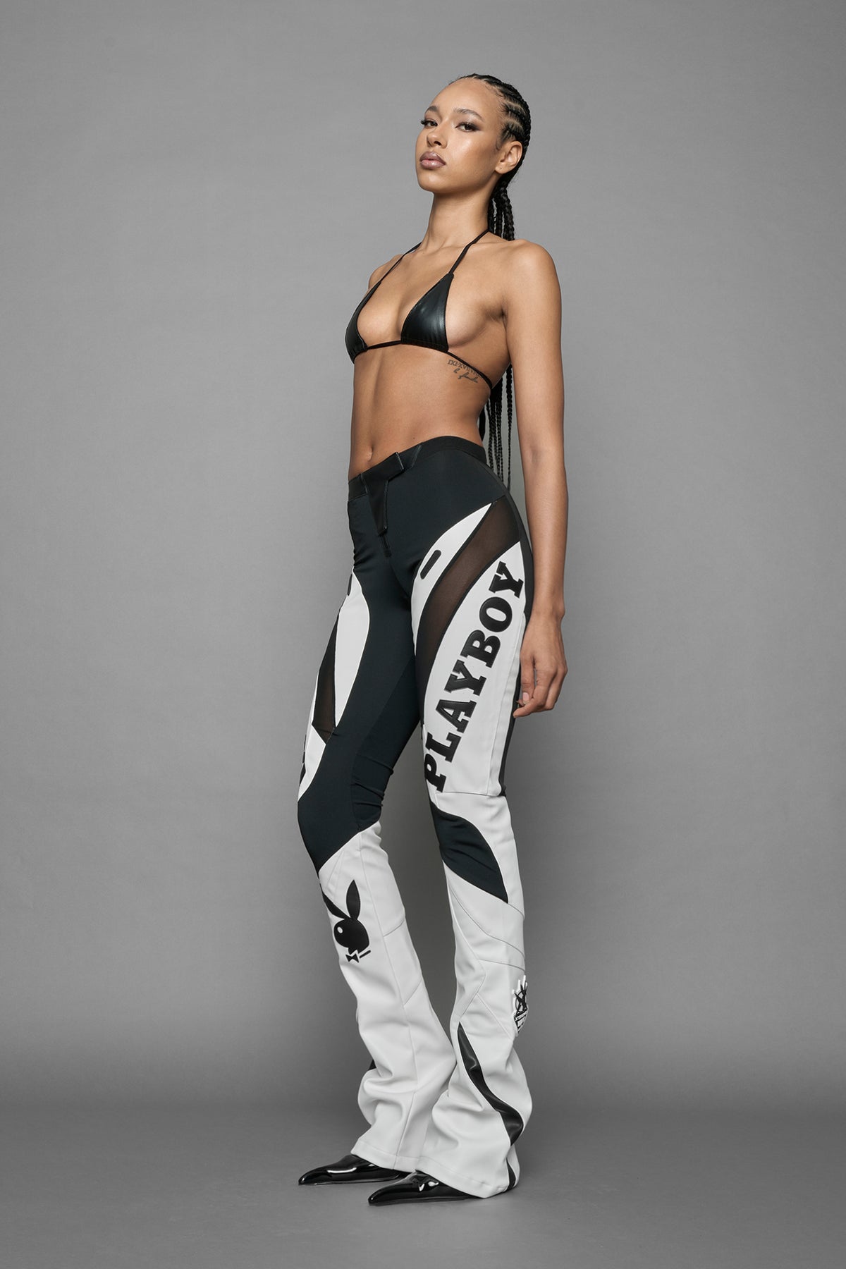 A female model wearing a white and black form fitted Pants with PU Leather Embroidery of 'Namilia', 'Playboy'. The pants have black Mesh fabric panels and are slightly flared from knee to hem