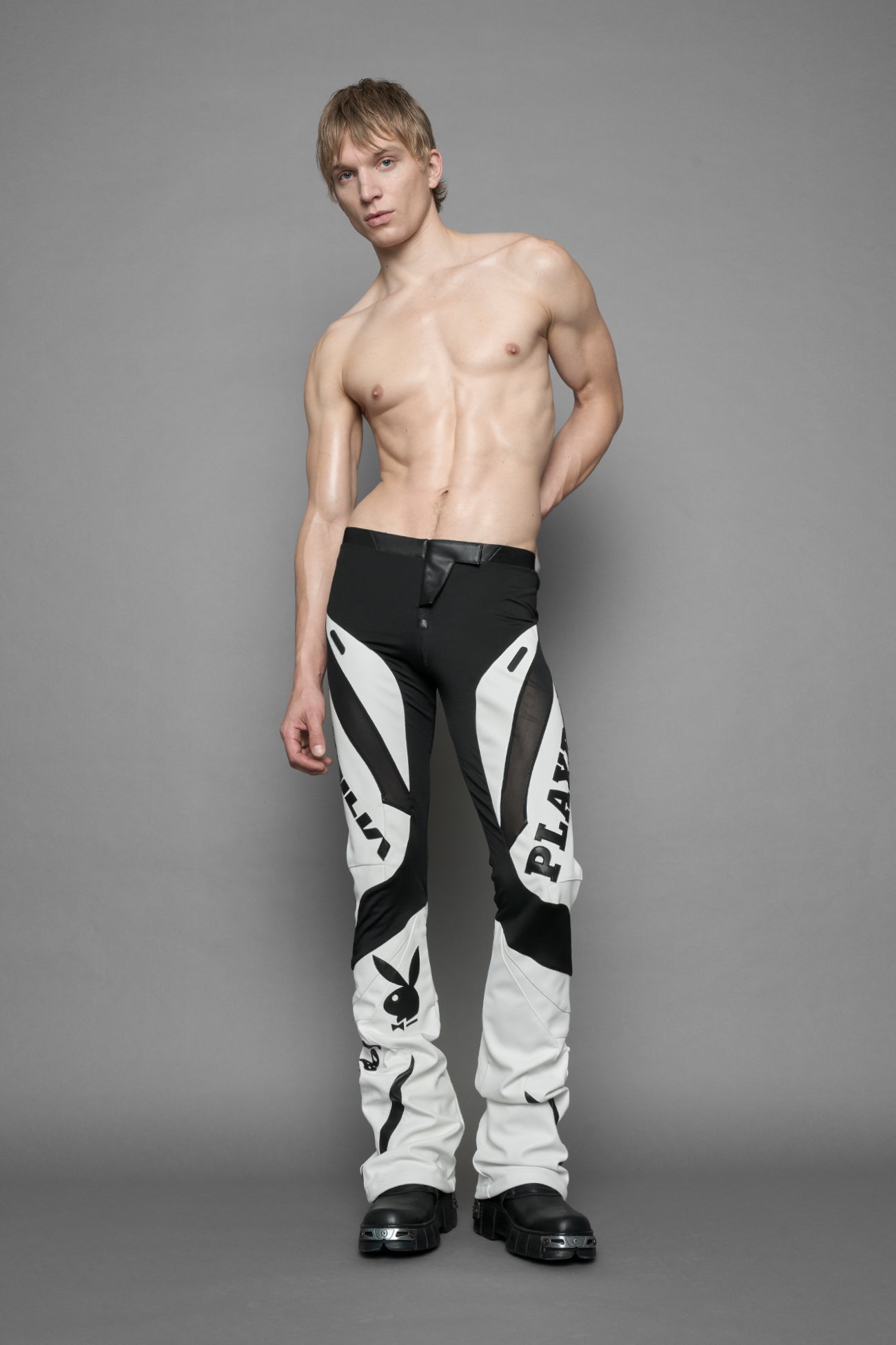 A female model wearing a white and black form fitted Pants with PU Leather Embroidery of 'Namilia', 'Playboy'. The pants have black Mesh fabric panels and are slightly flared from knee to hem