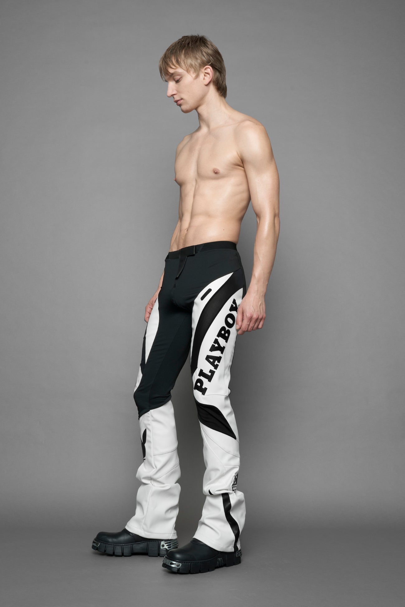 A female model wearing a white and black form fitted Pants with PU Leather Embroidery of 'Namilia', 'Playboy'. The pants have black Mesh fabric panels and are slightly flared from knee to hem