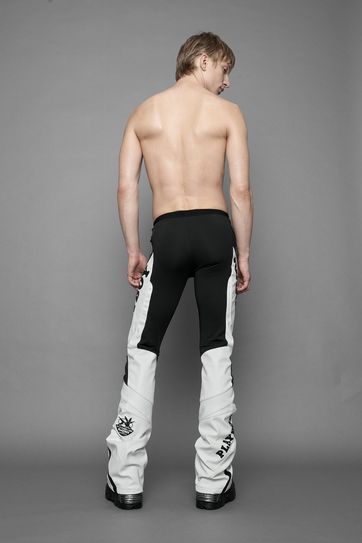 A female model wearing a white and black form fitted Pants with PU Leather Embroidery of 'Namilia', 'Playboy'. The pants have black Mesh fabric panels and are slightly flared from knee to hem