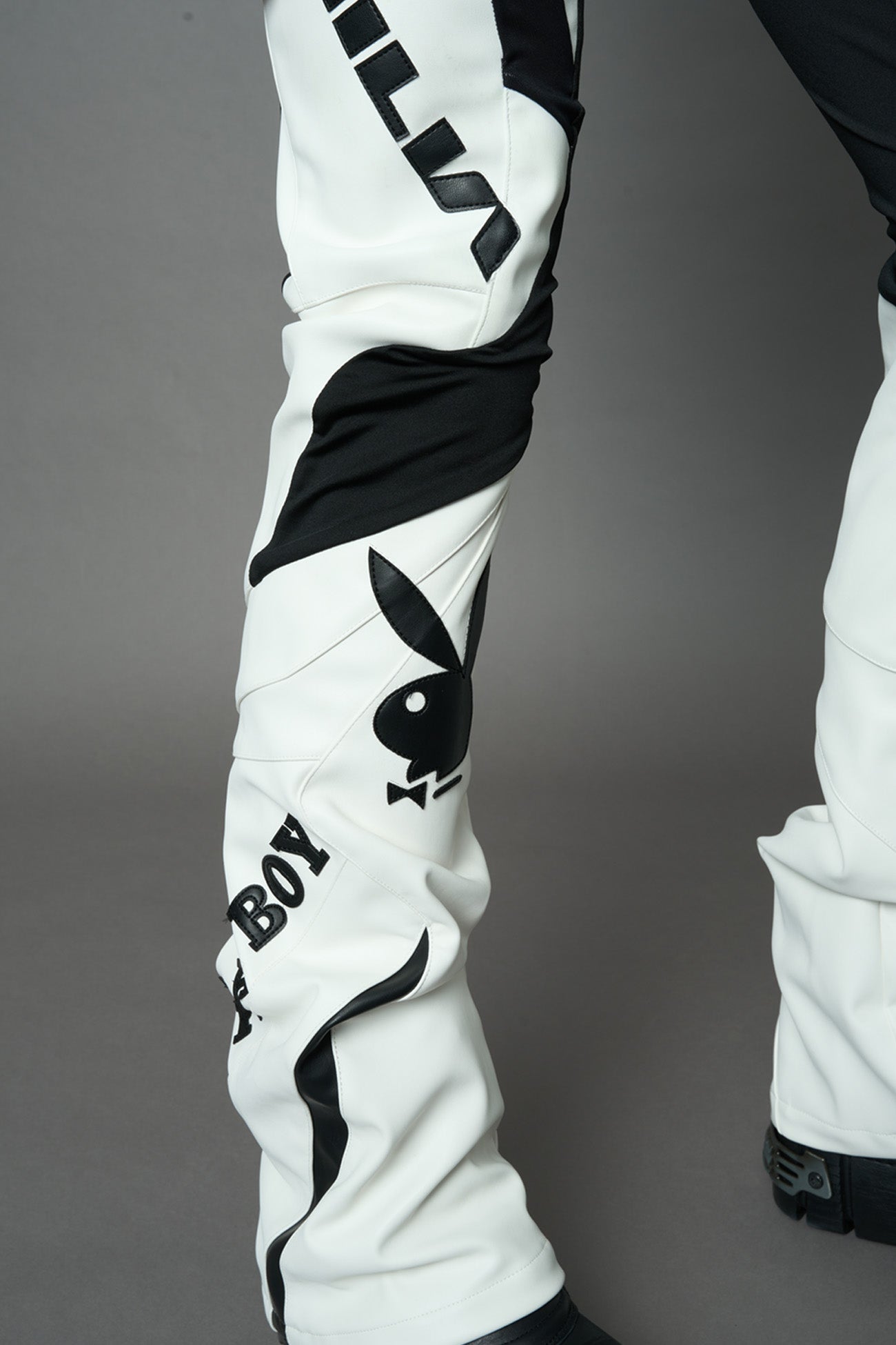 A female model wearing a white and black form fitted Pants with PU Leather Embroidery of 'Namilia', 'Playboy'. The pants have black Mesh fabric panels and are slightly flared from knee to hem
