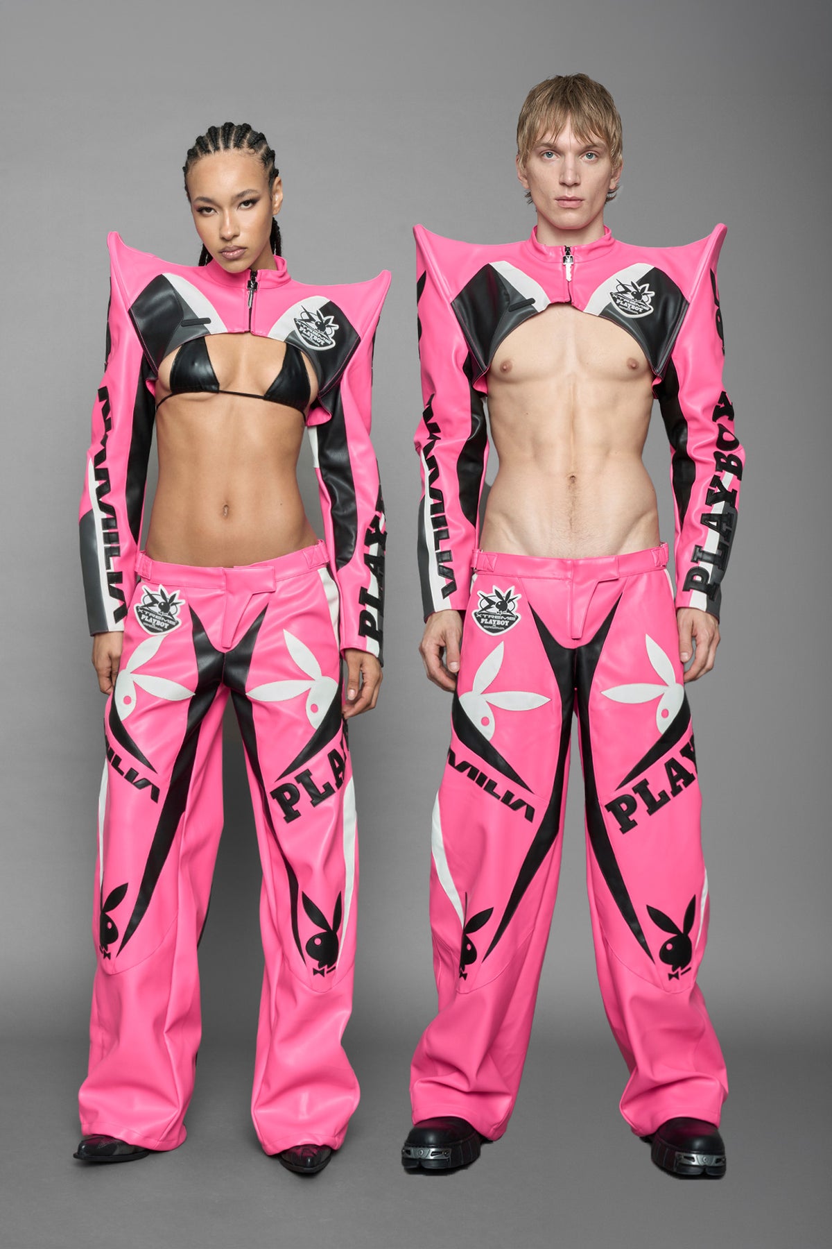  model wearing a vegan leather cropped jacket with spike silhouette shoulders in pink with black and white paneling. The jacket has PU leather embroidery of 'Namilia' and 'Playboy' with a key formed zipper pull and a rubbertag on the left breast.
