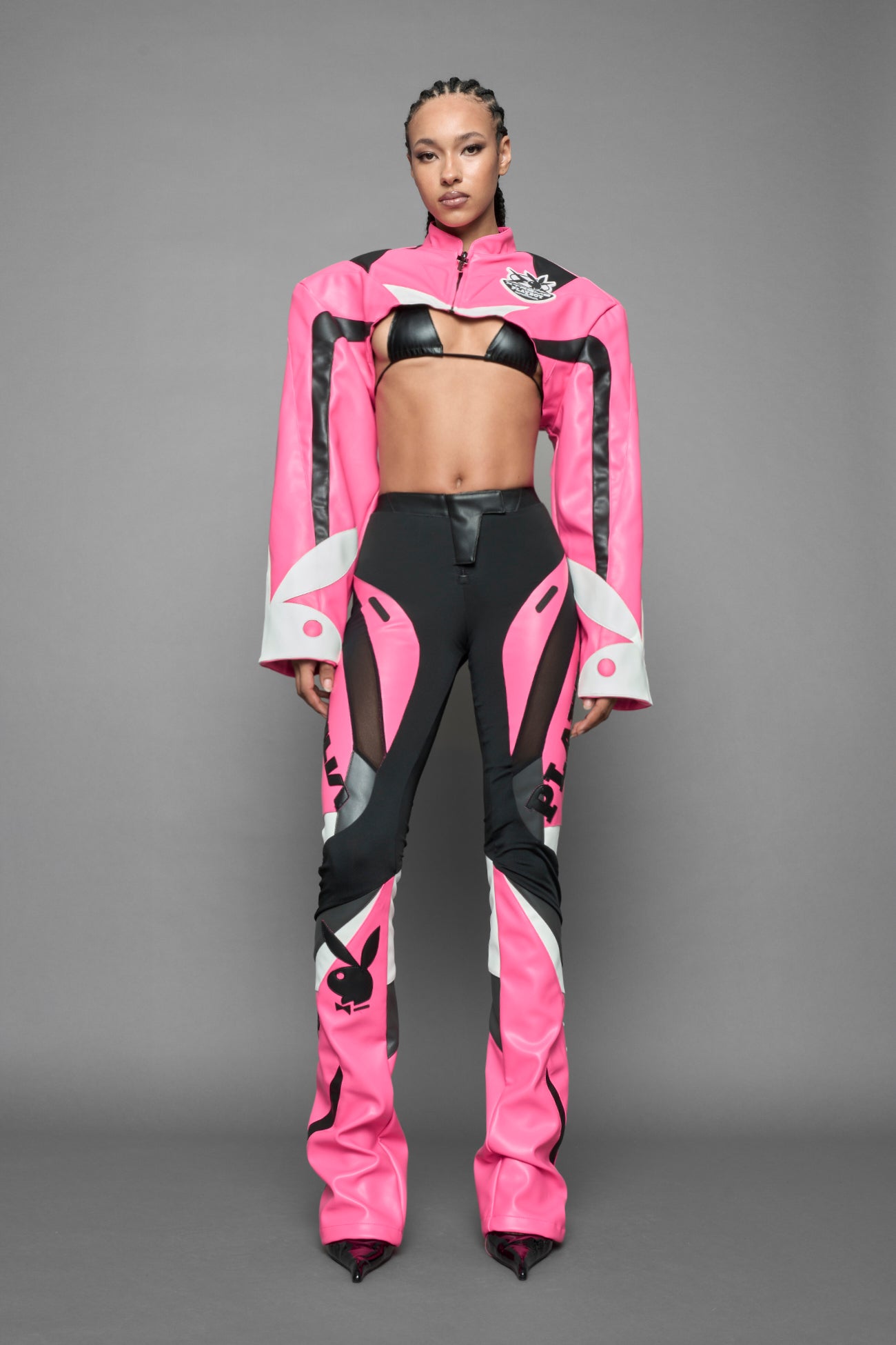 model wearing a vegan leather cropped jacket with broad shoulders in pink with black and white paneling. The jacket has PU leather embroidery of 'Namilia' and 'Playboy' with a key formed zipper pull and a rubbertag on the left breast.