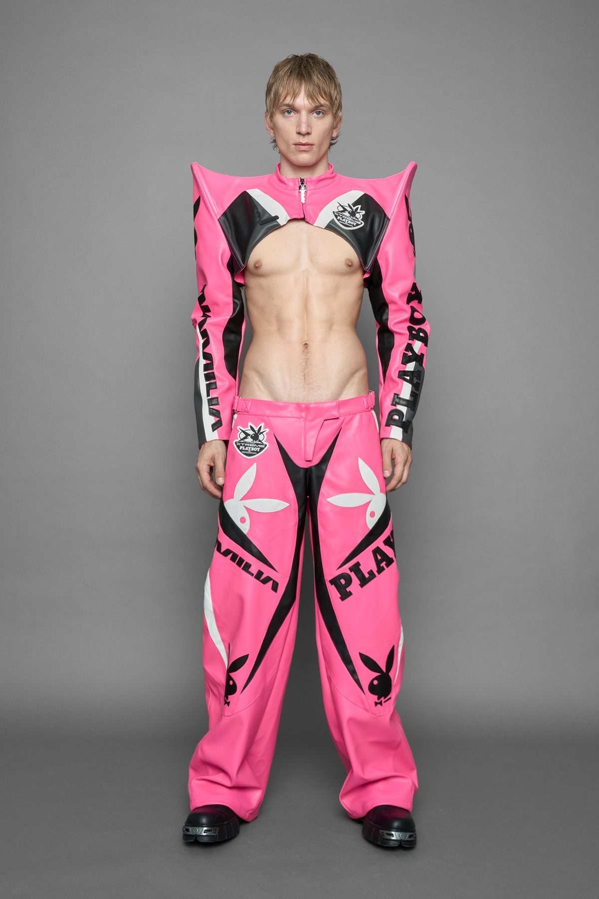  model wearing a vegan leather cropped jacket with spike silhouette shoulders in pink with black and white paneling. The jacket has PU leather embroidery of 'Namilia' and 'Playboy' with a key formed zipper pull and a rubbertag on the left breast.

