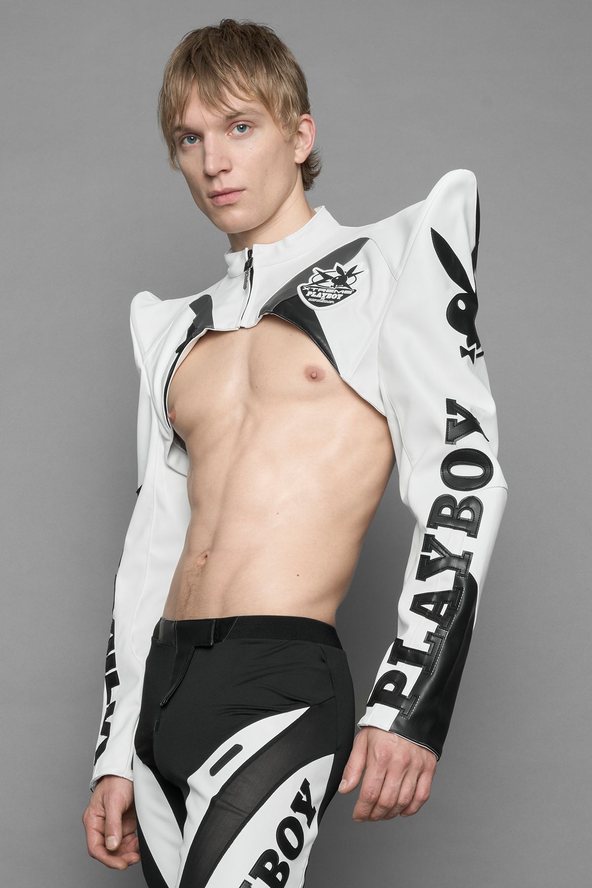 A model wearing a vegan leather cropped jacket with spike silhouette shoulders in white with black paneling. The jacket has PU leather embroidery of 'Namilia' and 'Playboy' with a key formed zipper pull and a rubbertag on the left breast.