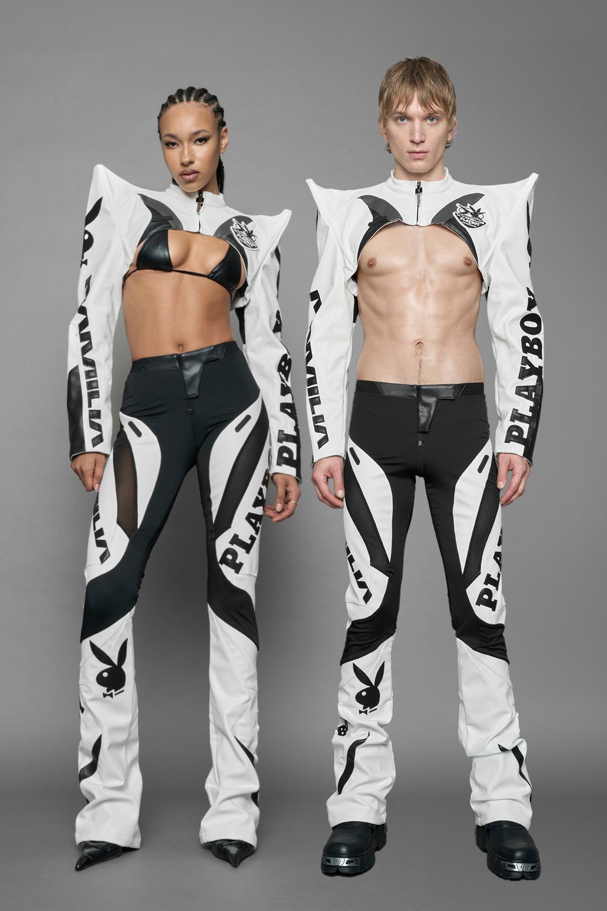 A female model wearing a vegan leather cropped jacket with spike silhouette shoulders in white with black paneling. The jacket has PU leather embroidery of 'Namilia' and 'Playboy' with a key formed zipper pull and a rubbertag on the left breast.