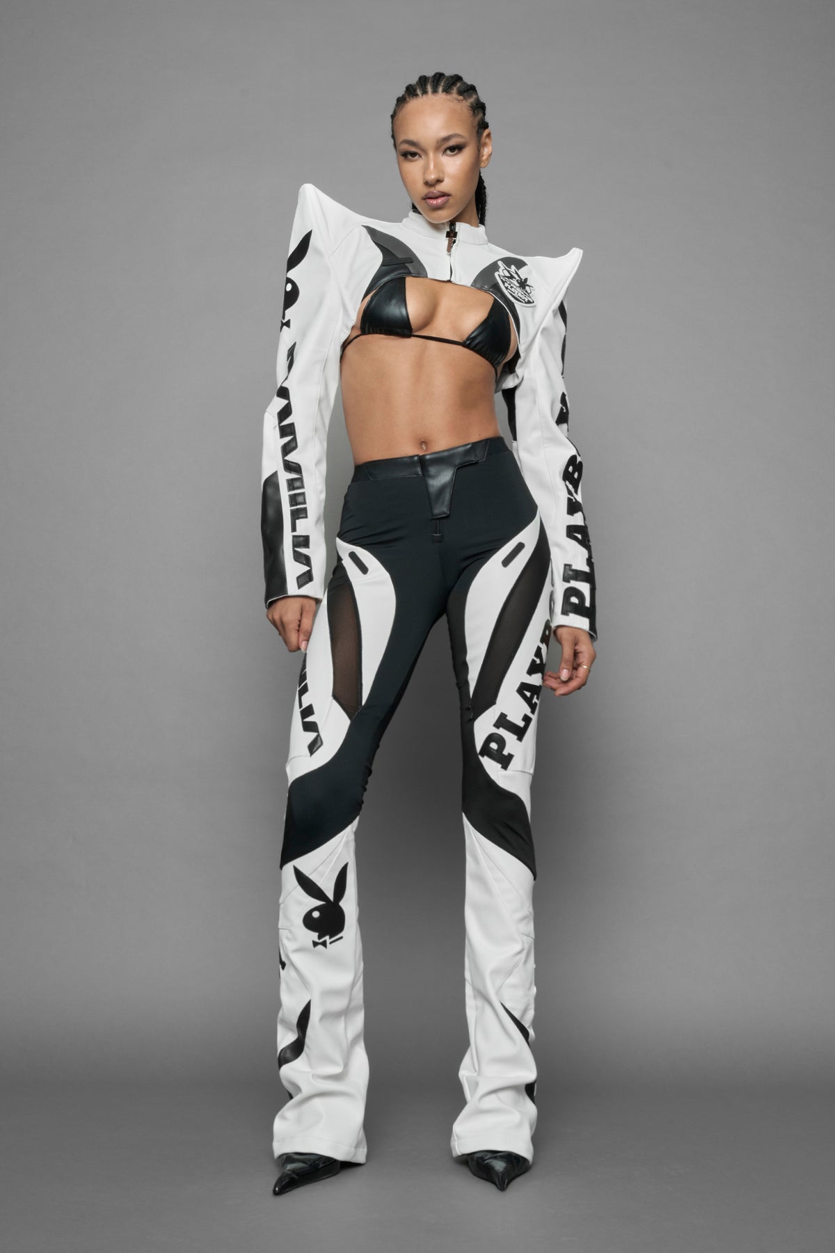 A female model wearing a vegan leather cropped jacket with spike silhouette shoulders in white with black paneling. The jacket has PU leather embroidery of 'Namilia' and 'Playboy' with a key formed zipper pull and a rubbertag on the left breast.