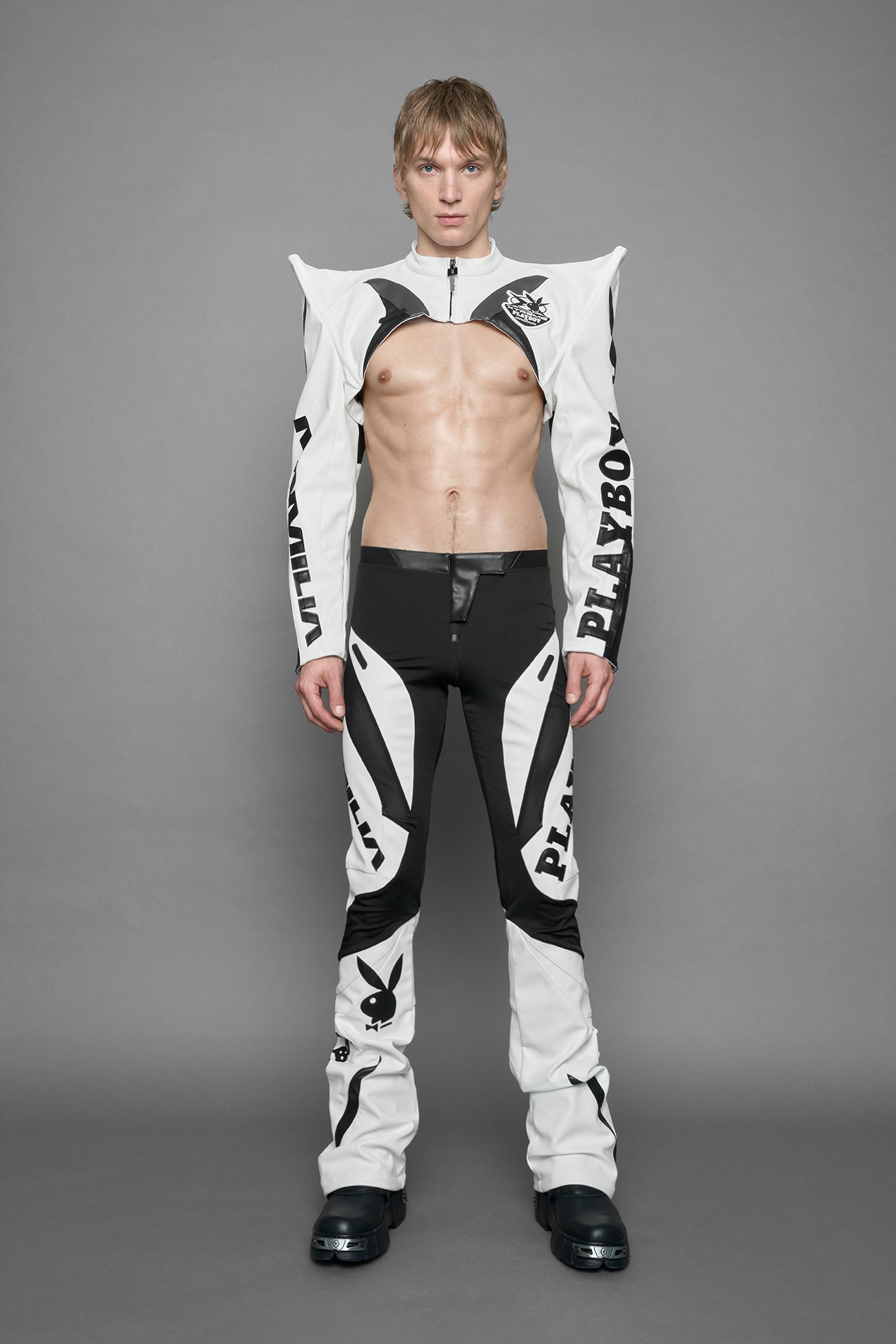 A model wearing a vegan leather cropped jacket with spike silhouette shoulders in white with black paneling. The jacket has PU leather embroidery of 'Namilia' and 'Playboy' with a key formed zipper pull and a rubbertag on the left breast.