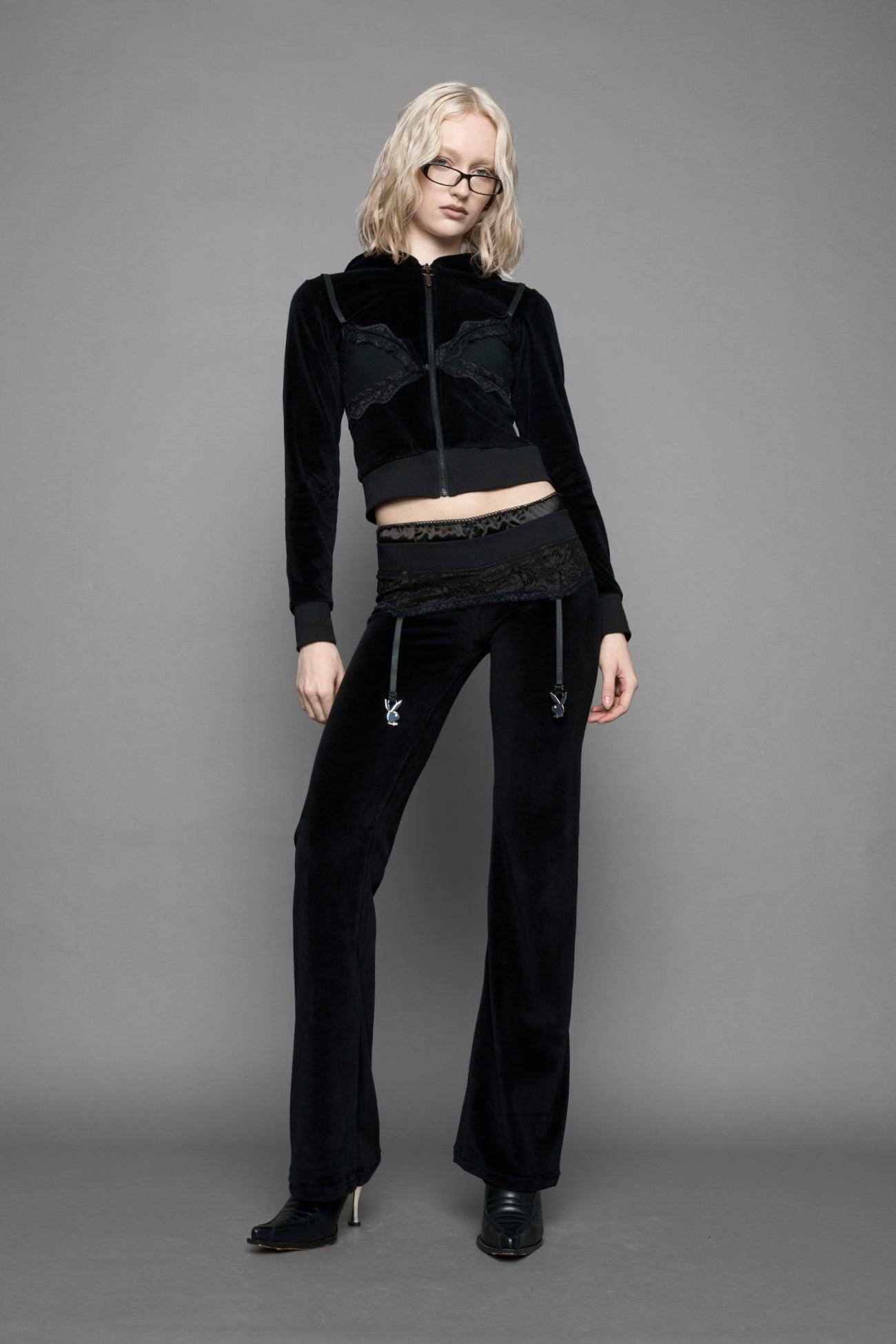 A female model wearing black velvet tracksuit pants with a lingerie black lace garter and suspenders with metal silver Bunnys 