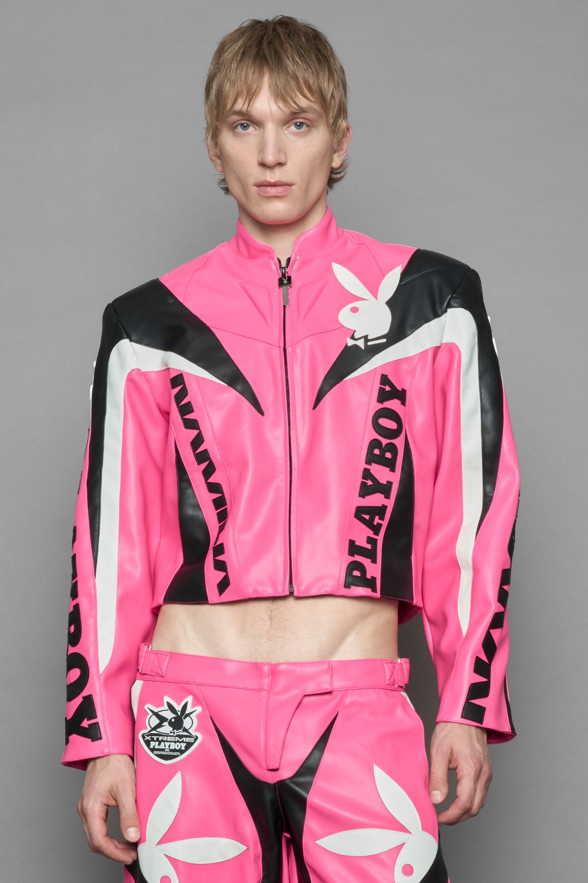 model wearing pink vegan leather zip-up jacket with black PU leather 'Namilia' and 'Playboy' embroidery and black and white paneling. On the left chest there is a white Playboy Bunny Embroidery.