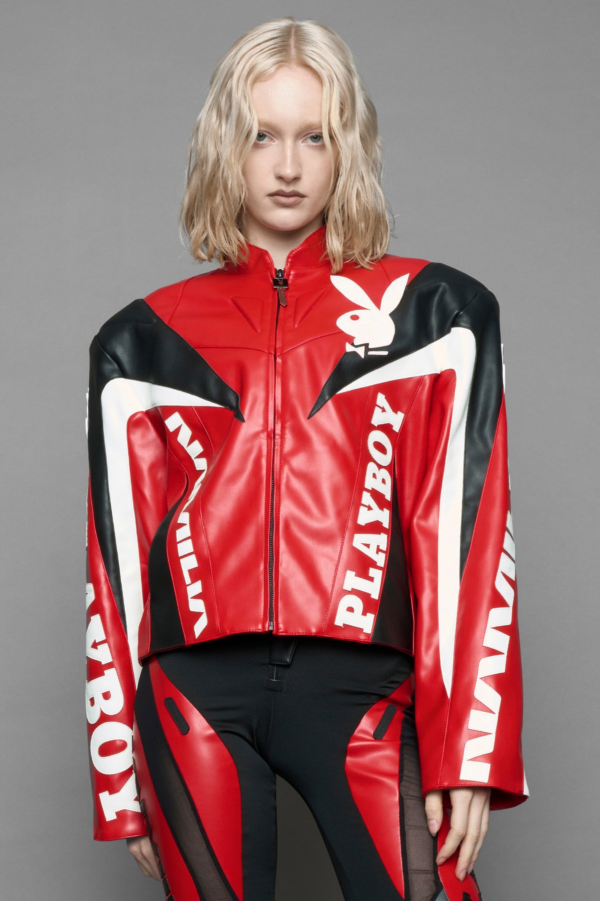 A female model wearing red vegan leather zip-up jacket with black PU leather 'Namilia' and 'Playboy' embroidery and black and white paneling. On the left chest there is a white Playboy Bunny Embroidery.