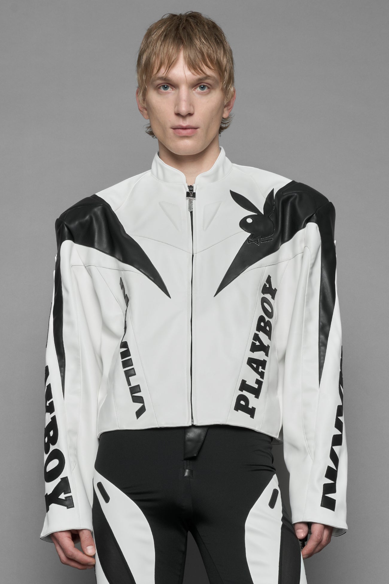 A male model wearing white vegan leather zip-up jacket with black PU leather 'Namilia' and 'Playboy' embroidery and black and white paneling. On the left chest there is a white Playboy Bunny Embroidery.