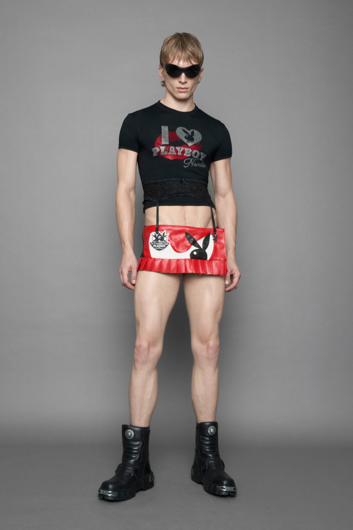 A male model wearing a red vegan leather wrap mini skirt with a black PU leather Bunny embroidery and a branded 'Playboy X Namilia' Rubbertag.