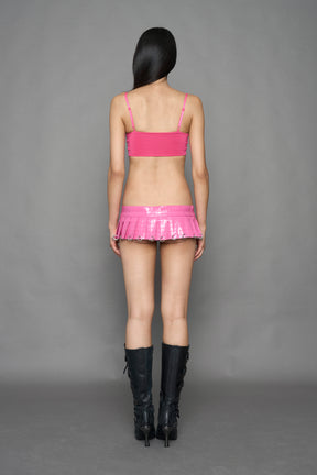 A model wearing a shiny pink two-piece outfit with a cropped strap top and matching pleated mini skirt, paired with tall black lace-up boots.