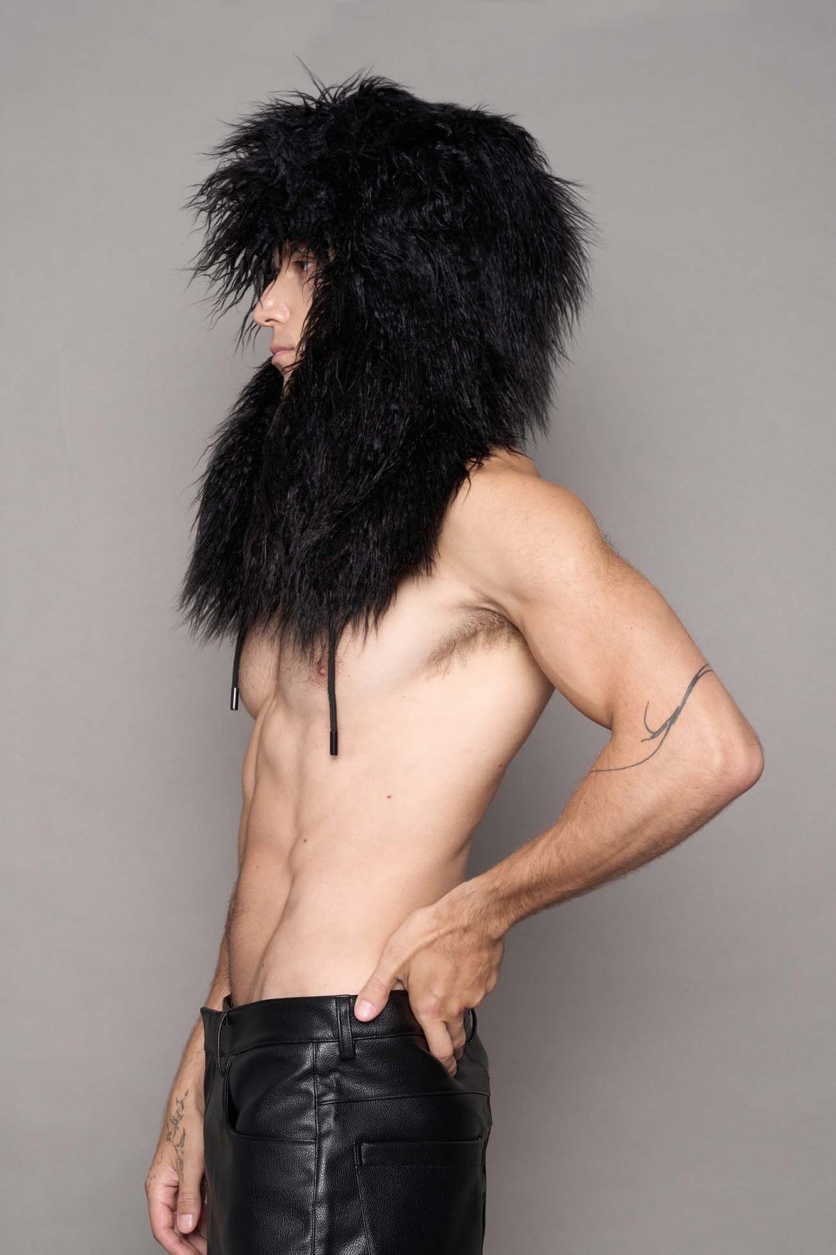 A shirtless man wearing a voluminous black faux fur oversized hat with earflaps and cords and leather pants posed against a plain gray studio background.