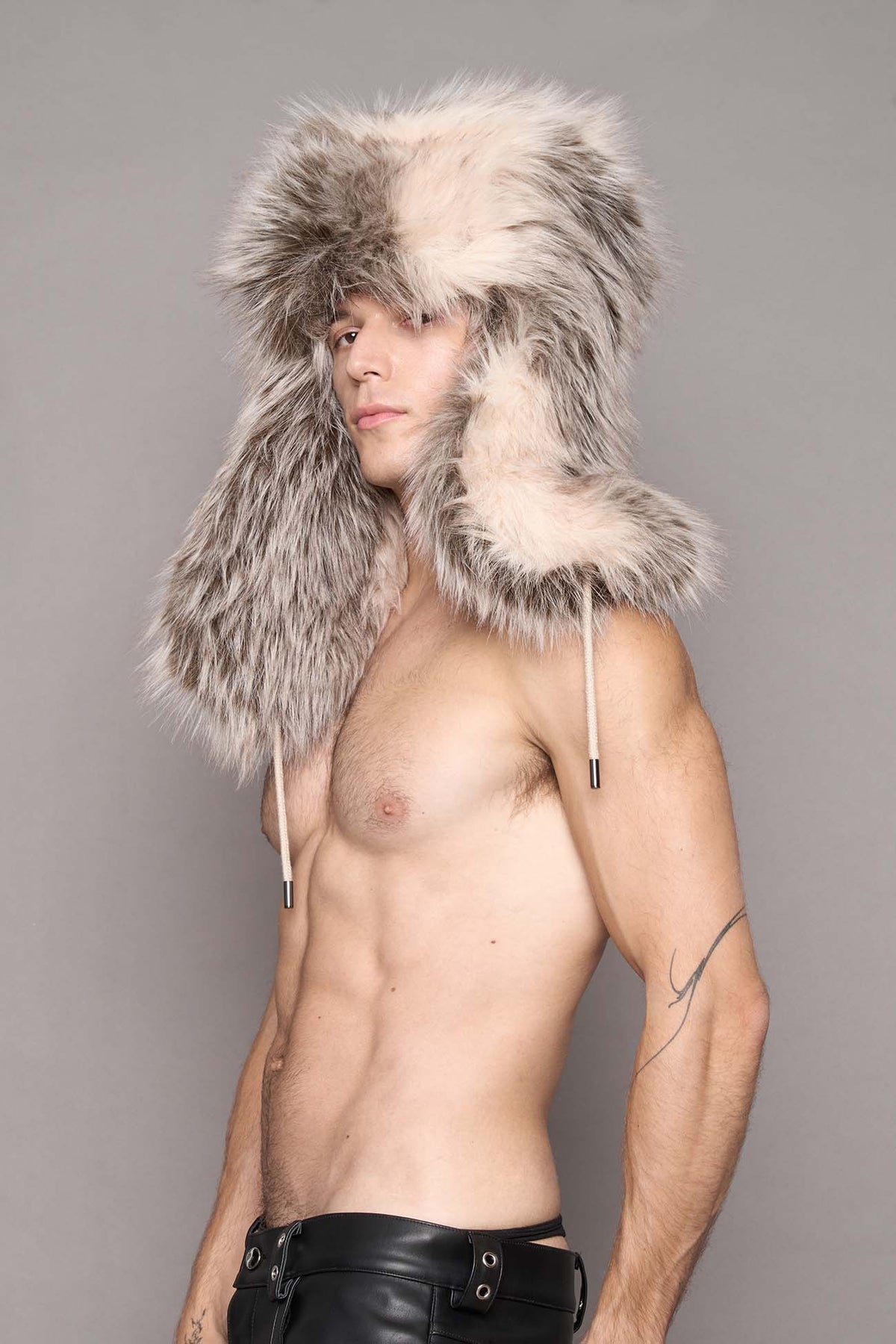 A shirtless man wearing a large, fluffy gray and cream fur ushanka hat, photographed against a solid gray studio background.