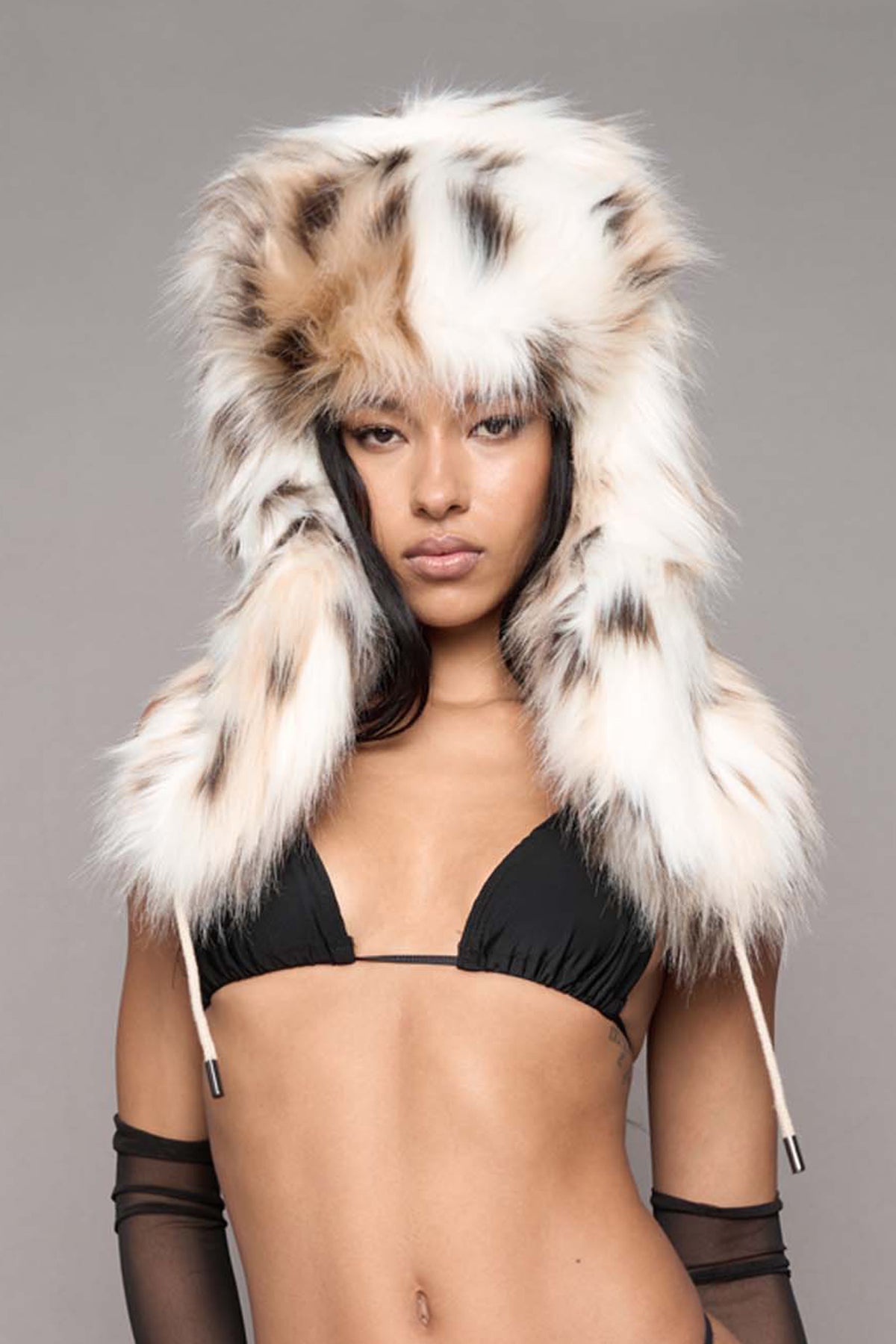 A model wearing a large, fluffy faux-fur hood with a black and cream spotted pattern, styled with black lingerie, sheer black arm sleeves, and black stockings against a solid gray background.