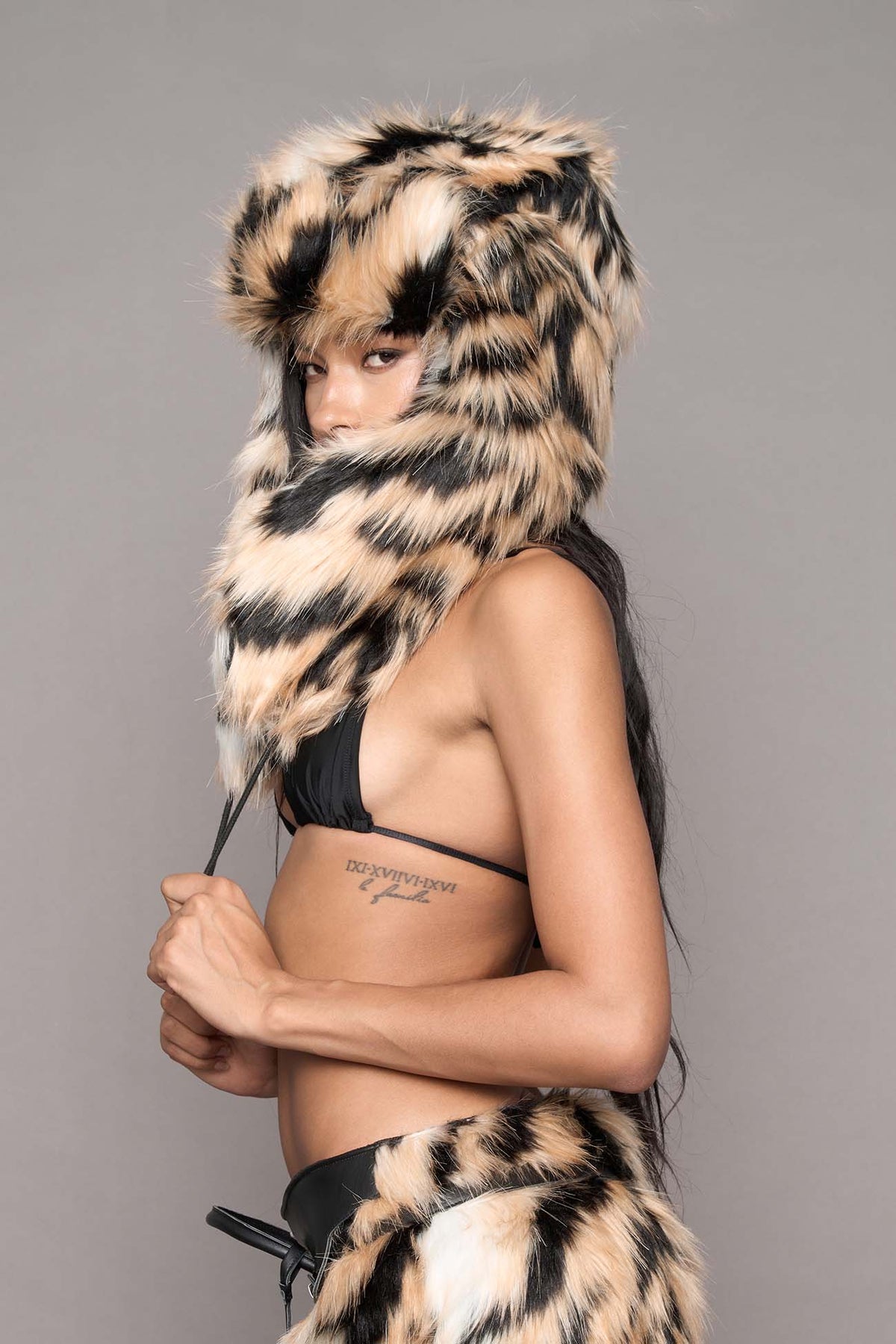 A model wearing a leopard-print faux fur hooded hat and matching leopard-print miniskirt , styled with a black bikini top against a solid gray studio background.