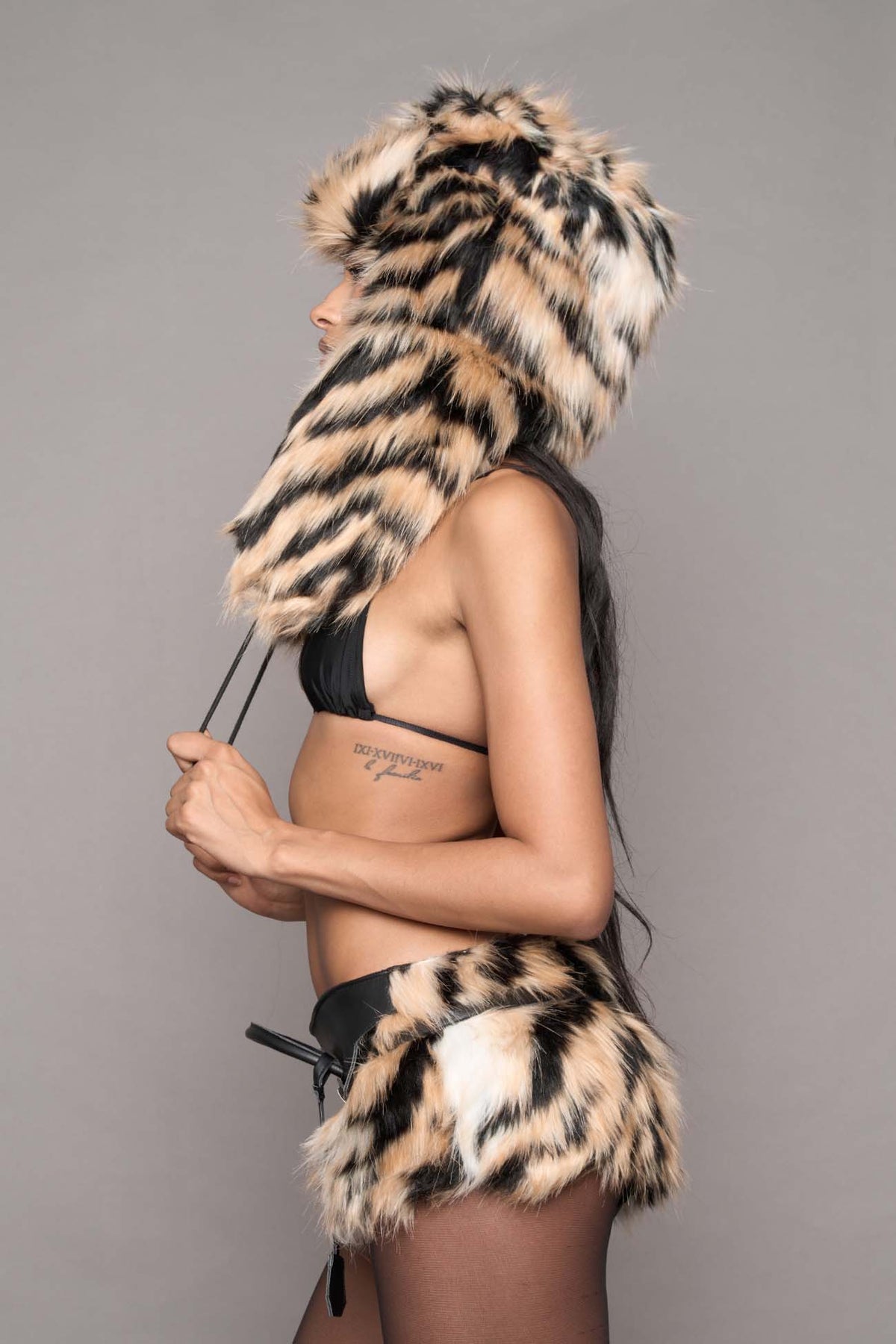 A model wearing a tiger faux fur hooded hat, a black bikini top, a black belt with a matching faux fur skirt, and sheer black tights, posed against a solid gray studio background.
